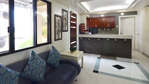A living room with a sofa and a coffee table, a kitchen with dark cabinets and a white countertop, and a large window with blinds. - The Windy Ridge Inn (Tagaytay)