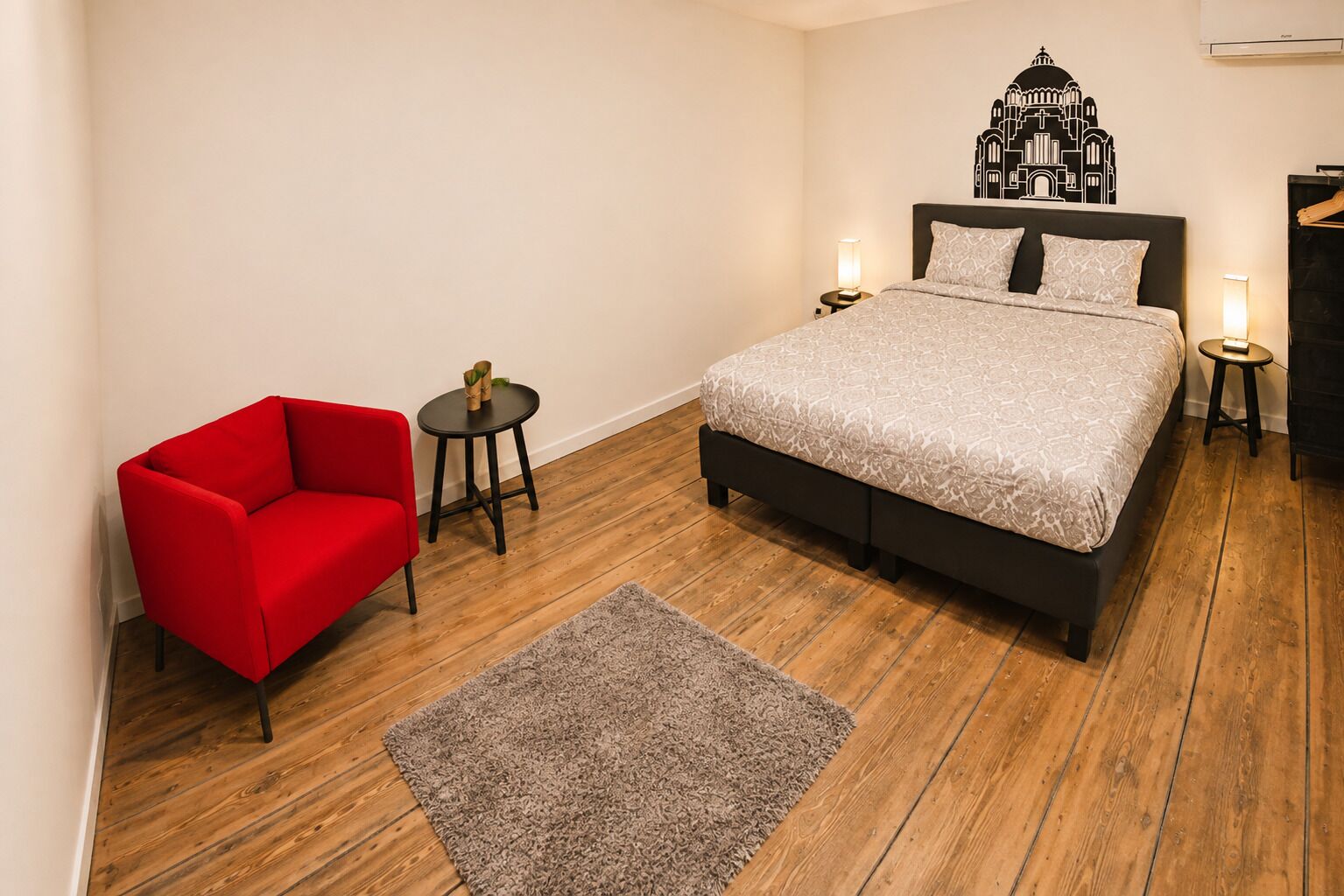 A bedroom with a bed, a red chair, a small table, and a decorative wall piece.