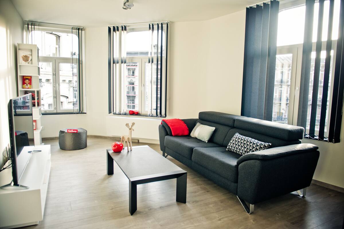 A modern living room with a grey sofa, a black coffee table, and a red ottoman. There are two windows with curtains, and a city view is visible through the windows.