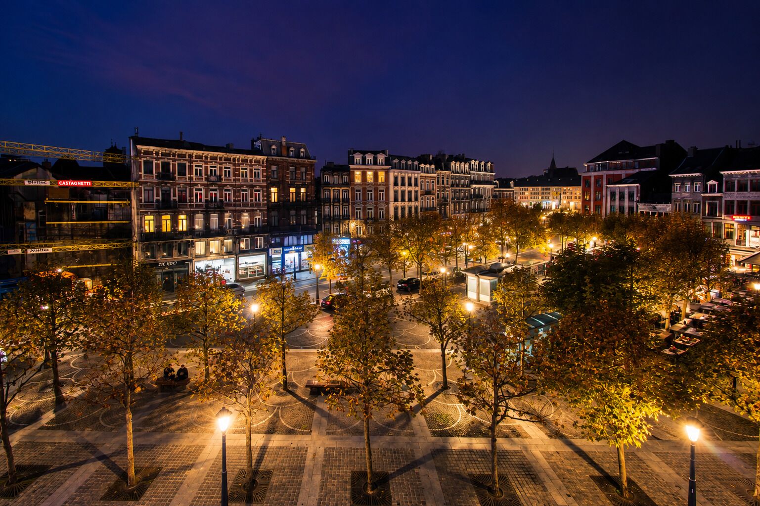 A nighttime cityscape with illuminated buildings and a tree-lined square.