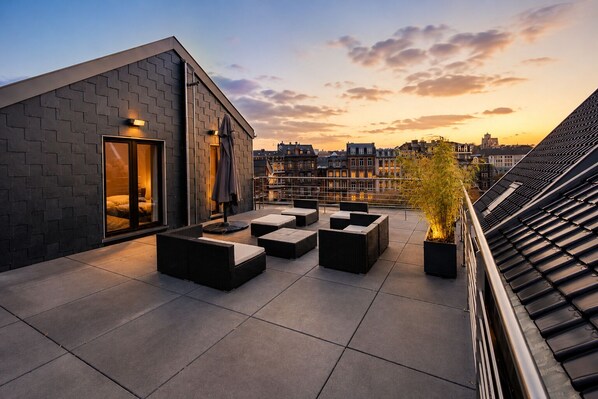 A rooftop terrace with outdoor seating, a potted plant, and a view of the city skyline at sunset. - Les Suites de Nanesse (Liege)