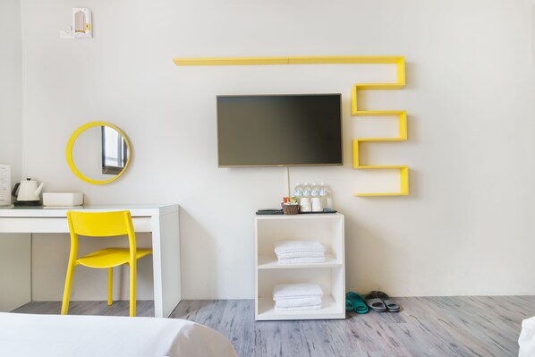 A modern hotel room with a white desk, yellow chair, a flat-screen TV mounted on the wall, a mirror, a shelf with toiletries, and a pair of slippers on the floor.