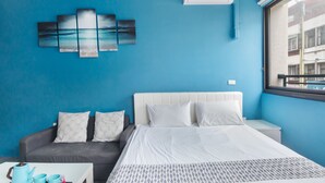 A hotel room with a bed, a gray sofa, a white coffee table with cups and a kettle, and a blue wall with a painting. - MUNA Hostel (Hualien City)