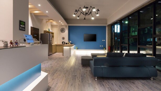 A modern living room with a blue accent wall, a large flat-screen TV, a dark blue sofa, and a kitchen area in the background. - MUNA Hostel (Hualien City)