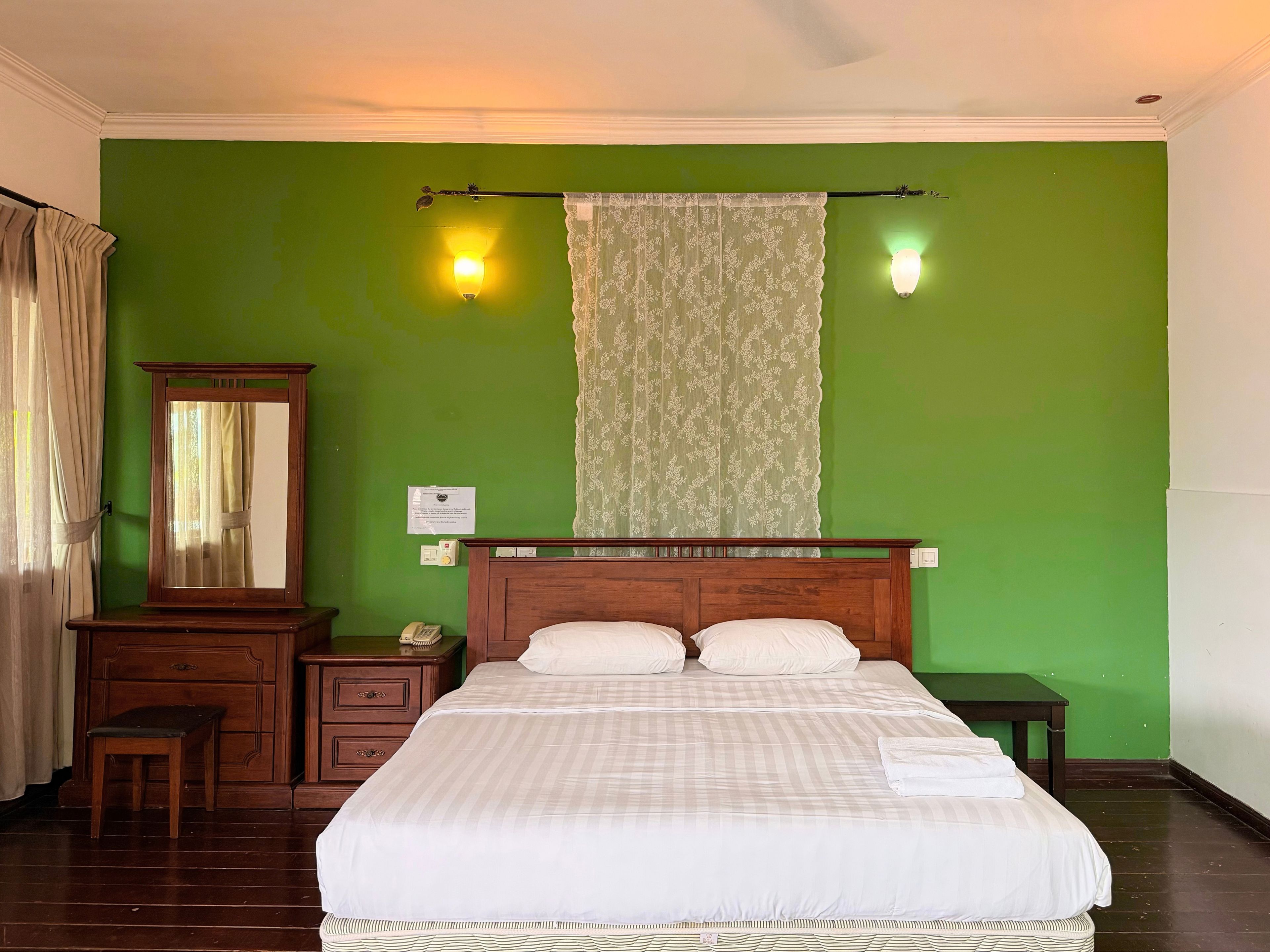 A hotel room with a bed, a wooden dresser, a mirror, a nightstand, a lamp, and a window with curtains.
