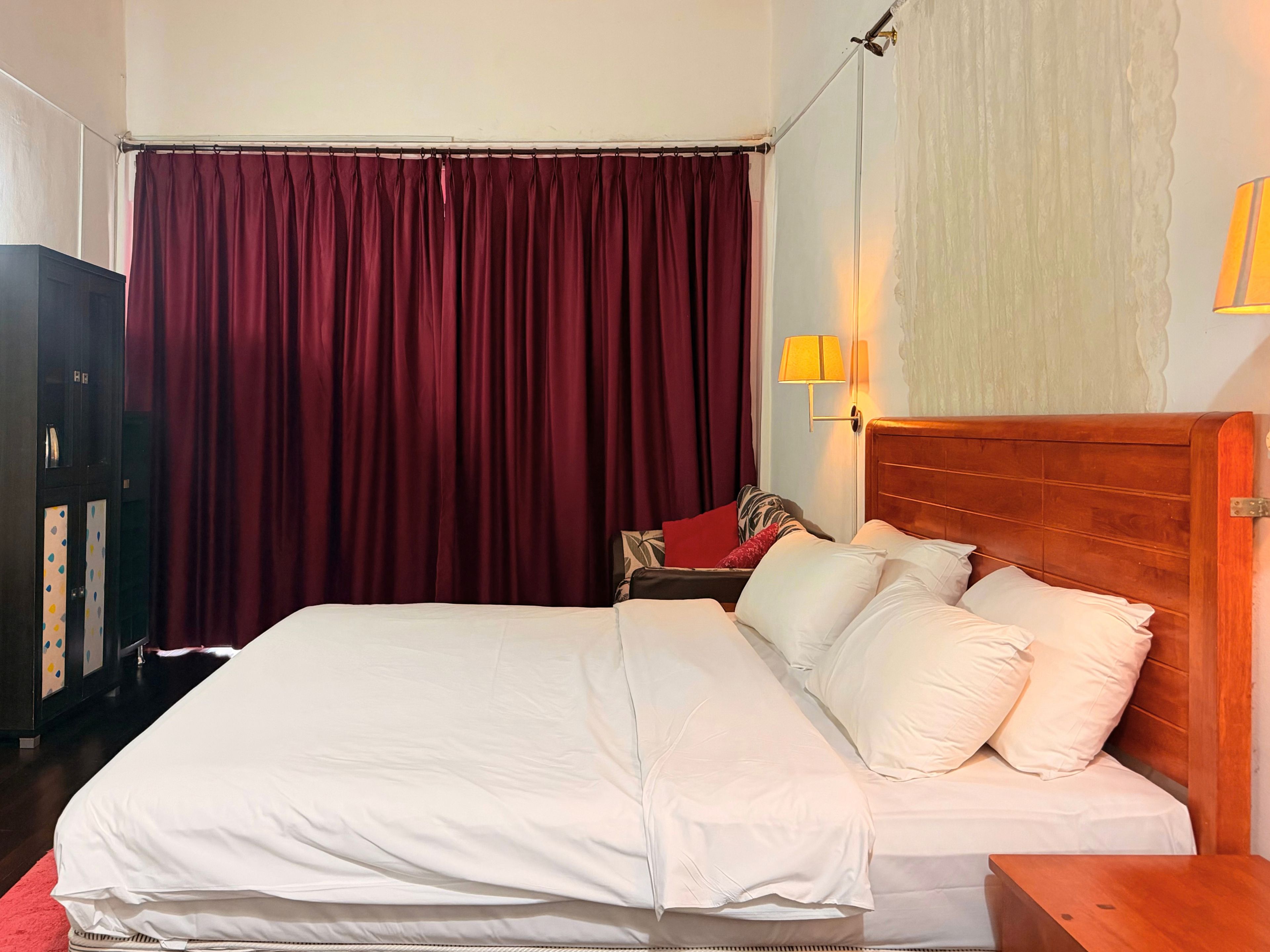 A hotel room with a large bed, a wooden headboard, a nightstand, a lamp, a wardrobe, and a curtain.