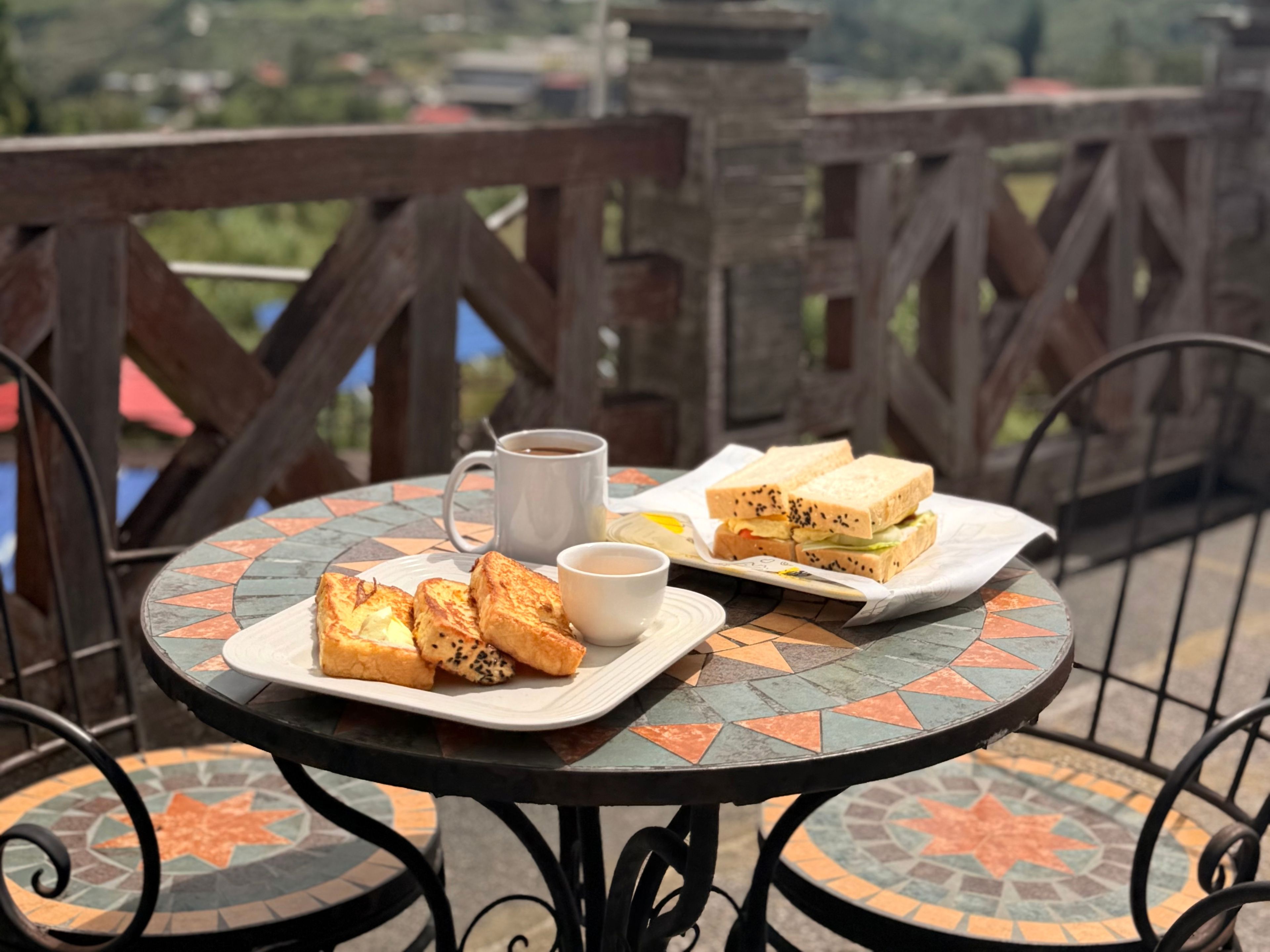 A table with a meal consisting of sandwiches, toast, and a cup of coffee, set on a tiled patio with a view of a bridge and greenery.