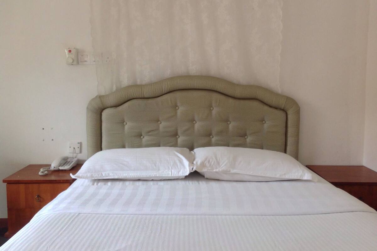 A neatly made bed with a striped headboard, a wooden nightstand with a telephone, and a plain wall with a light switch.