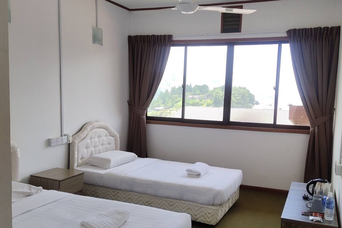 A hotel room with two beds, a ceiling fan, a window with curtains, a small table with a kettle, and a green trash bin.