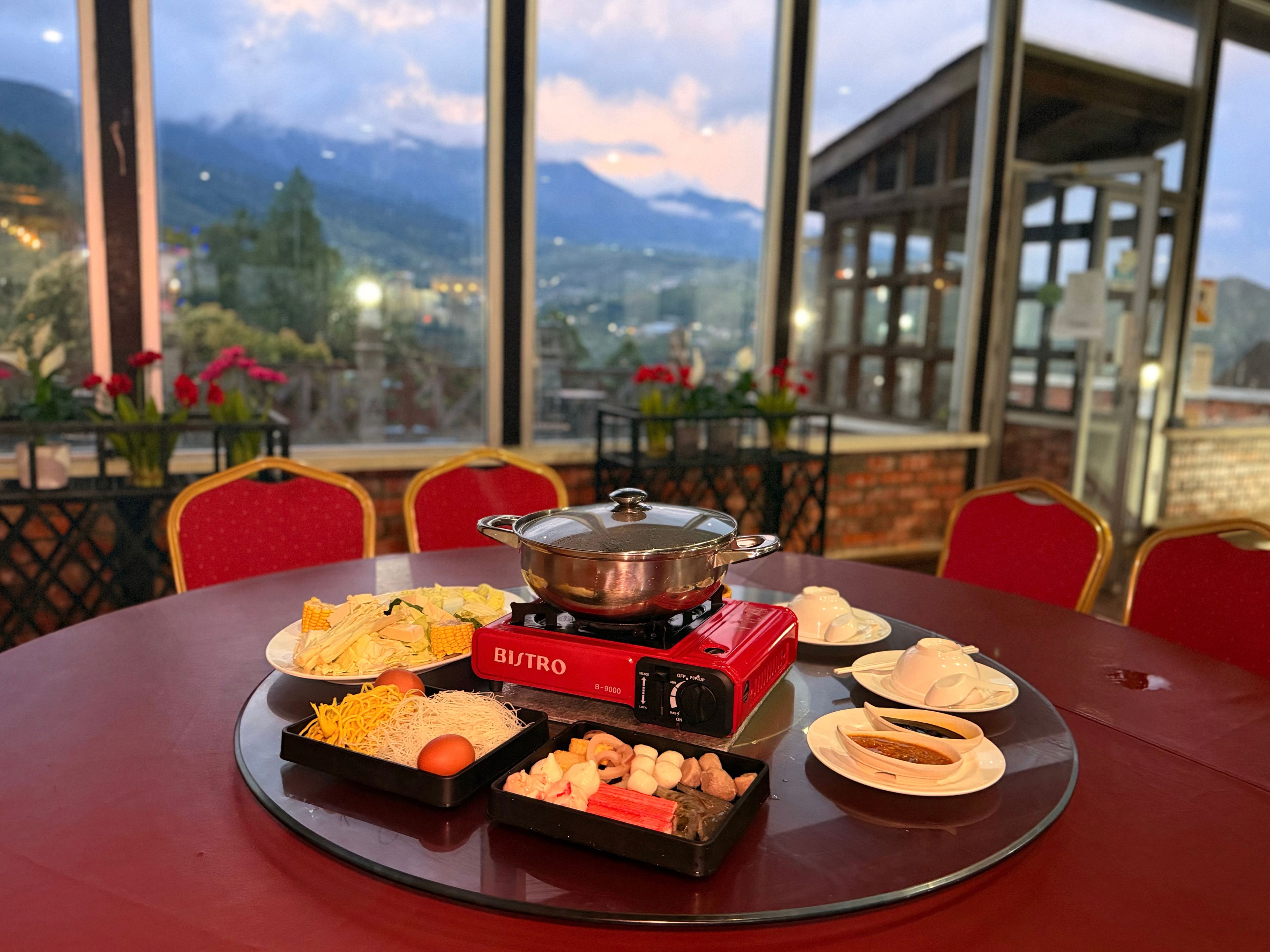 A dining table with a BISTRO brand portable stove, various dishes, and a scenic mountain view through large windows.