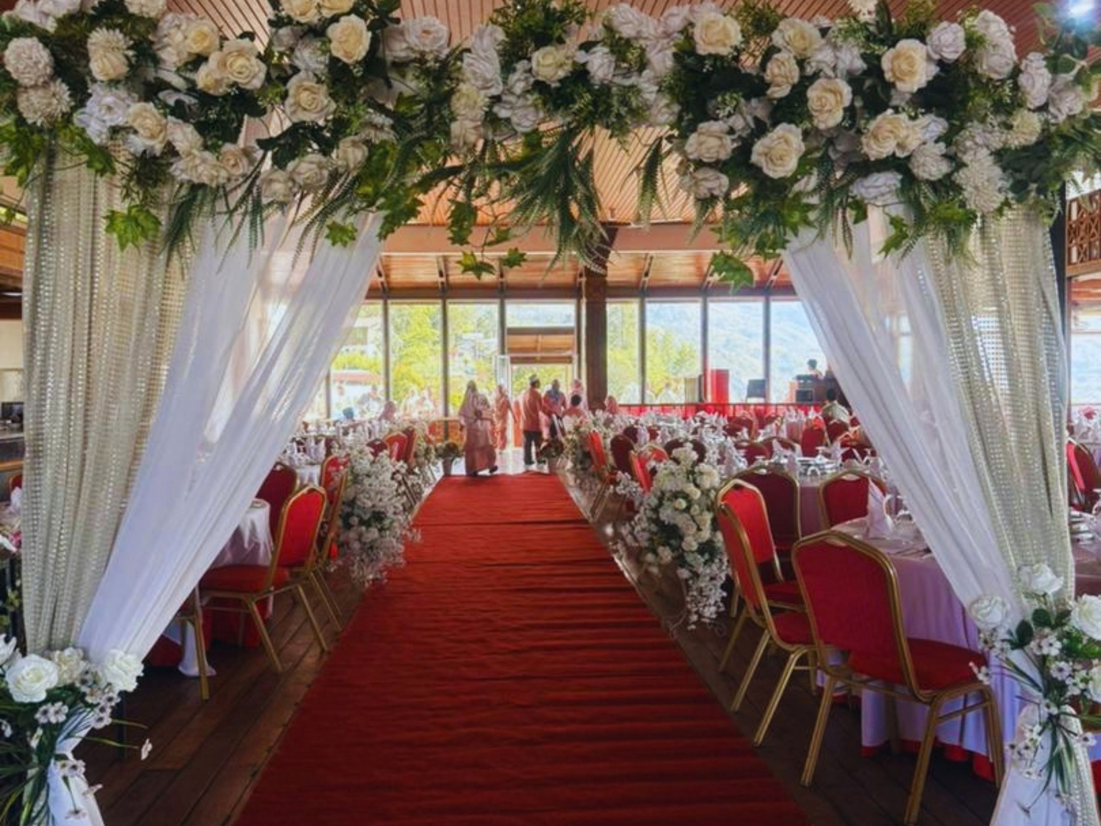 An indoor venue with a red carpet aisle, white floral decorations, and red chairs.
