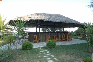 Un área de bar al aire libre con techo de paja, taburetes de madera y piso de concreto.
