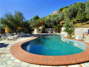 Una piscina de borde curvo, rodeada de sillas de descanso y sombrillas, con árboles y una colina de fondo.