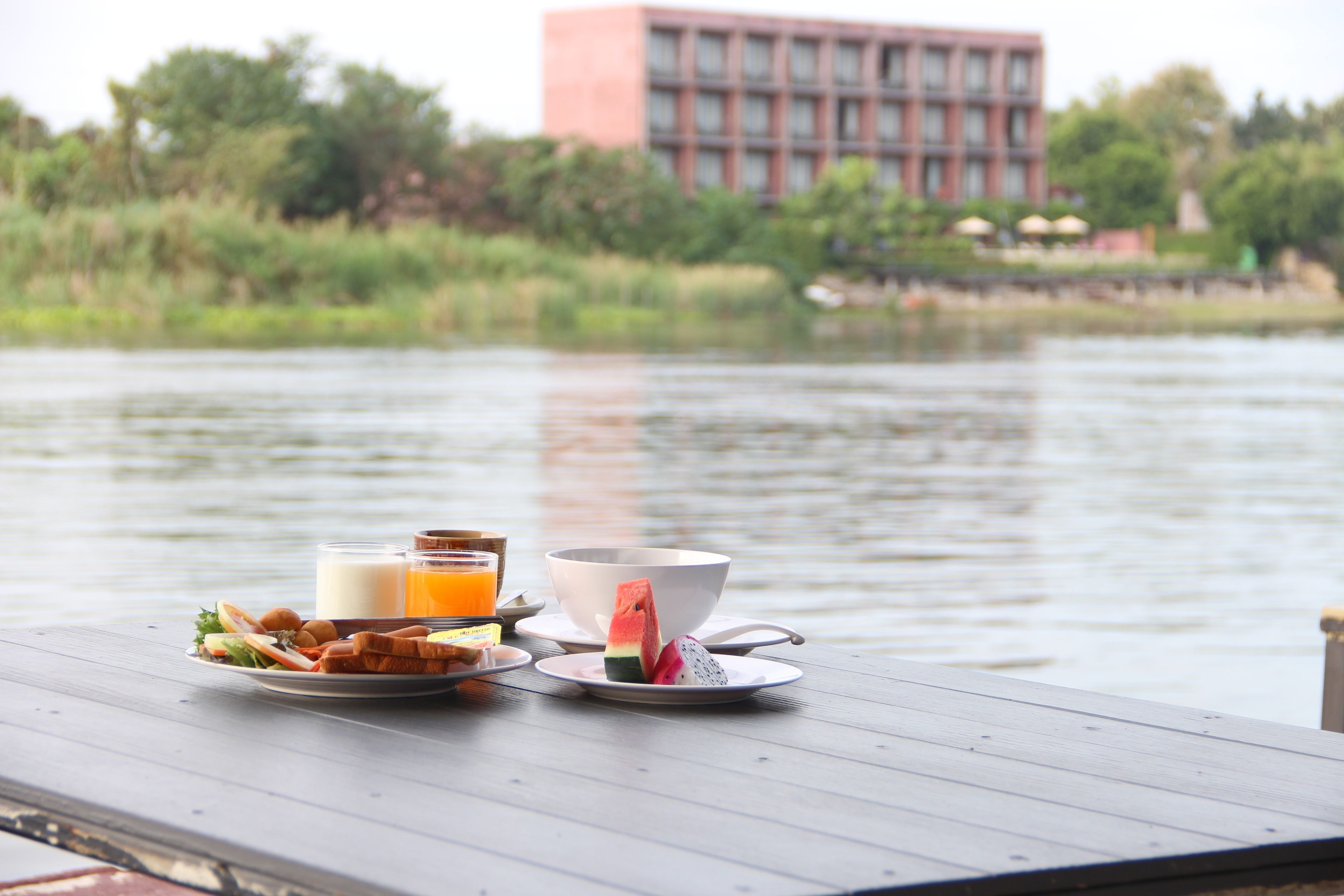 A breakfast table with food and drinks by a lake, with a building in the background.