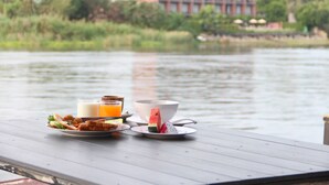 A breakfast table with food and drinks by a lake, with a building in the background.