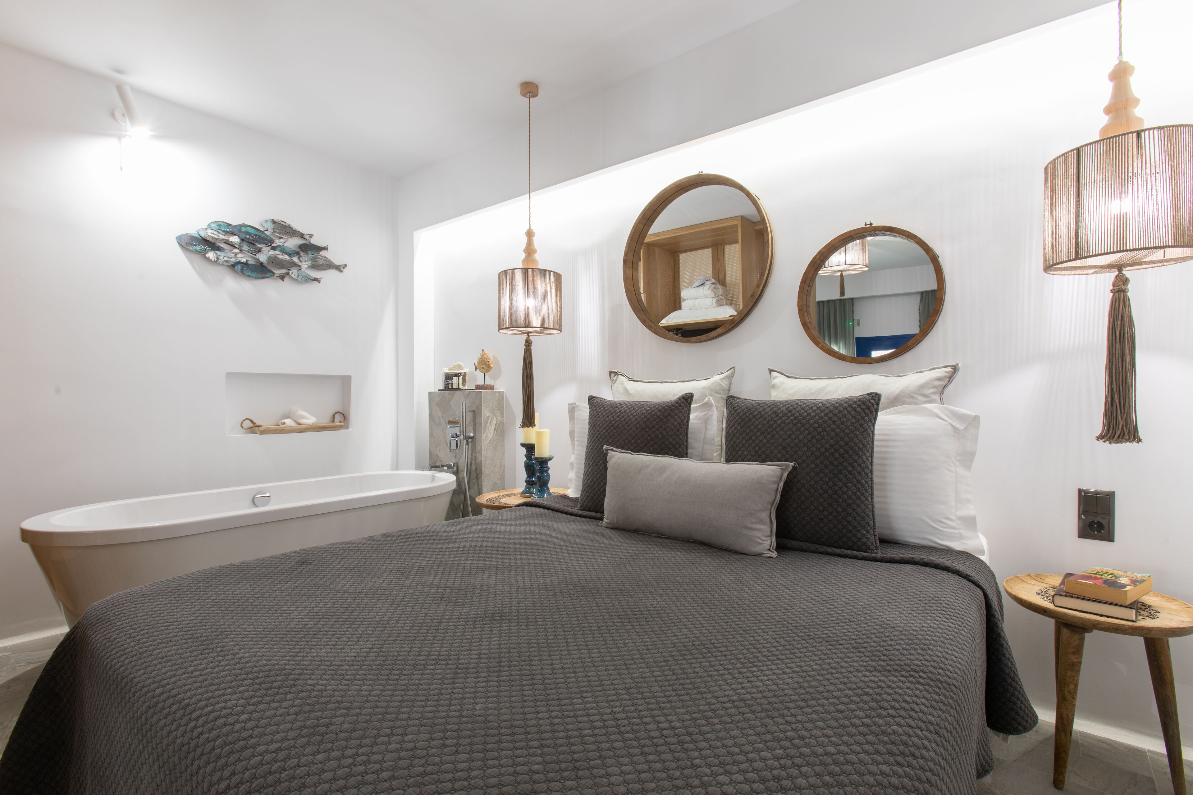 A bedroom with a large bed, two bedside tables, a round mirror, a hanging lamp, a wall-mounted shelf, and a bathtub.