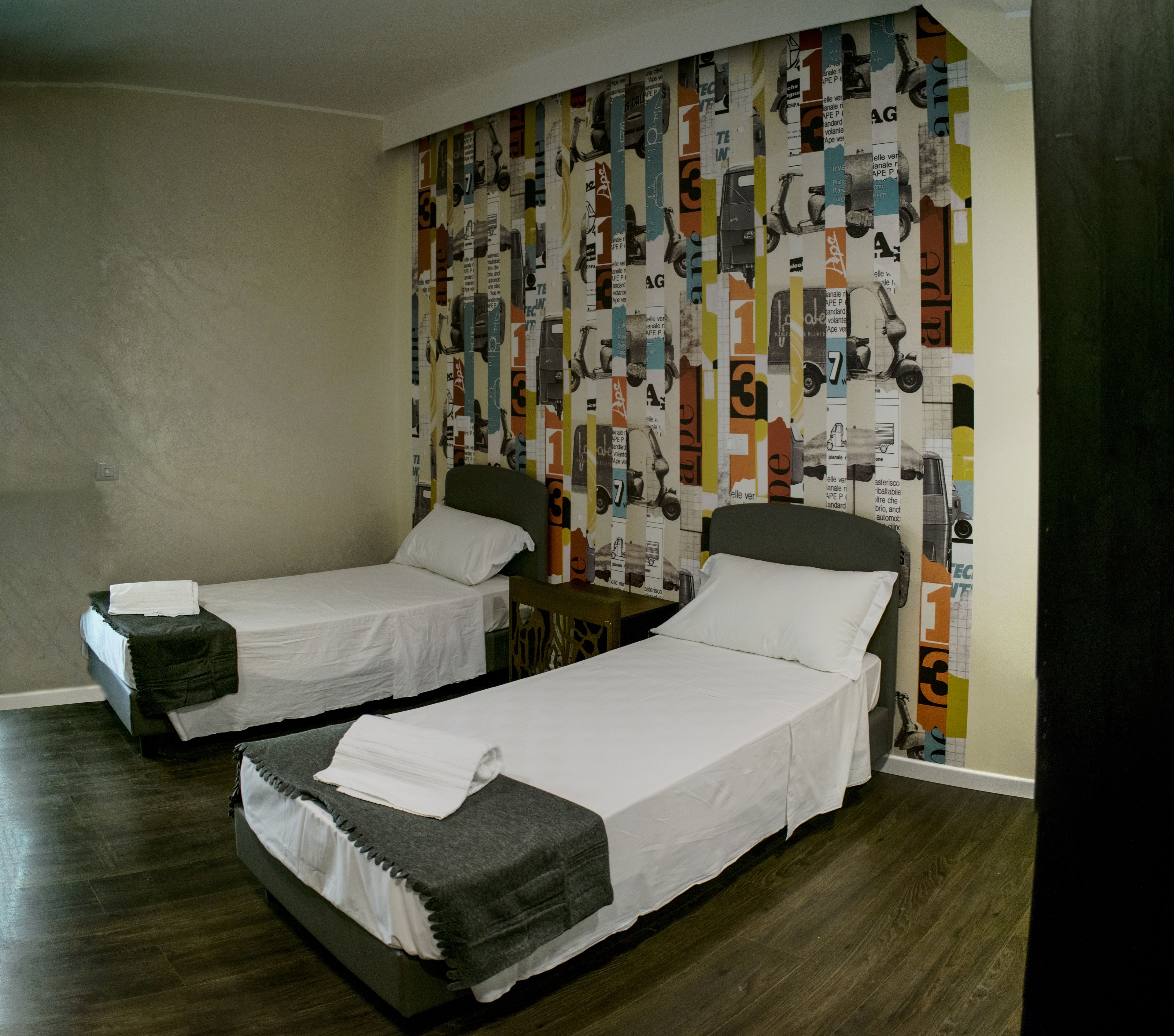 Camera d'albergo con due letti singoli, pavimento in legno e parete colorata con diversi motivi.