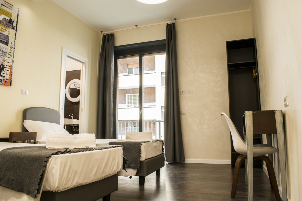 A hotel room with two beds, a chair, a wardrobe, and a view of the outside balcony.
