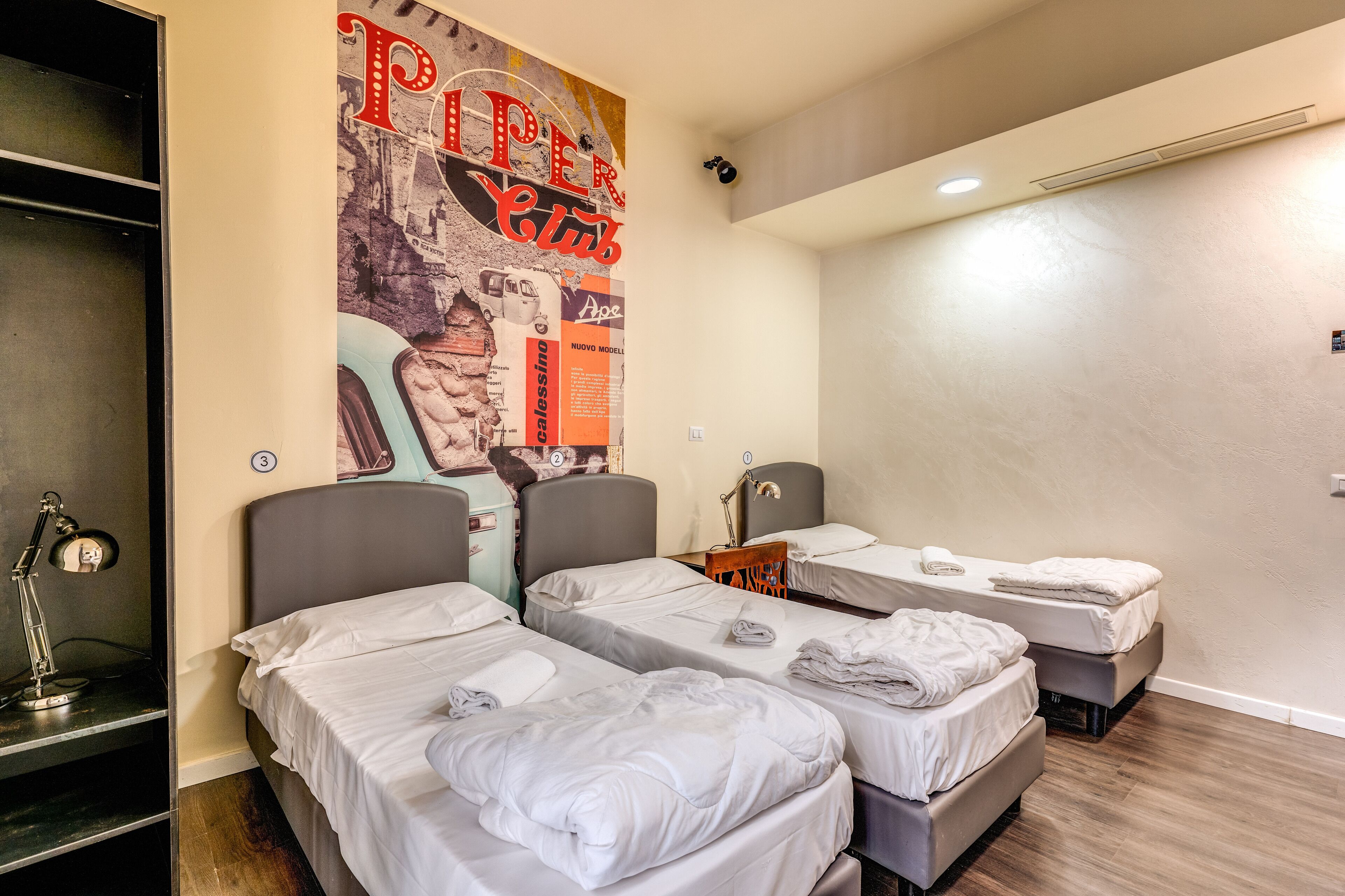 Habitación de hotel con tres camas, un escritorio con lámpara y un cartel grande en la pared.