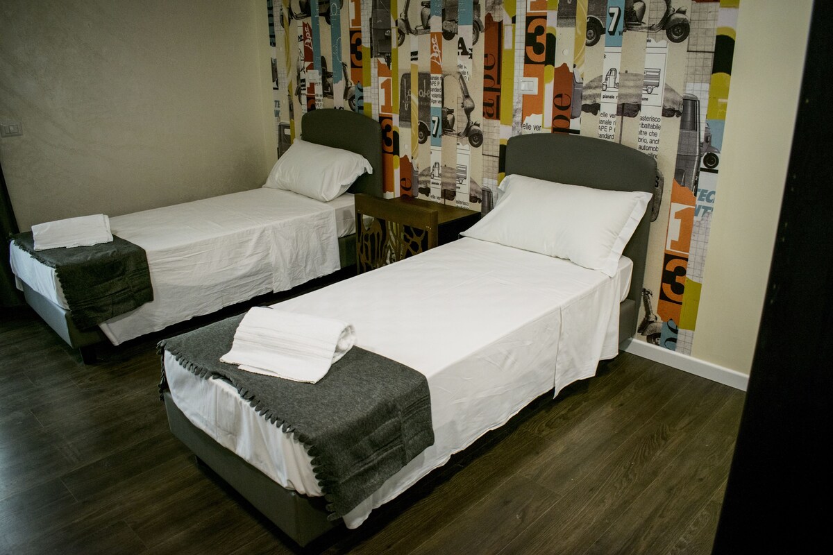 A room with two single beds, each with a pillow and a blanket. The room has a wooden floor and a wall with a colorful, patterned wallpaper.