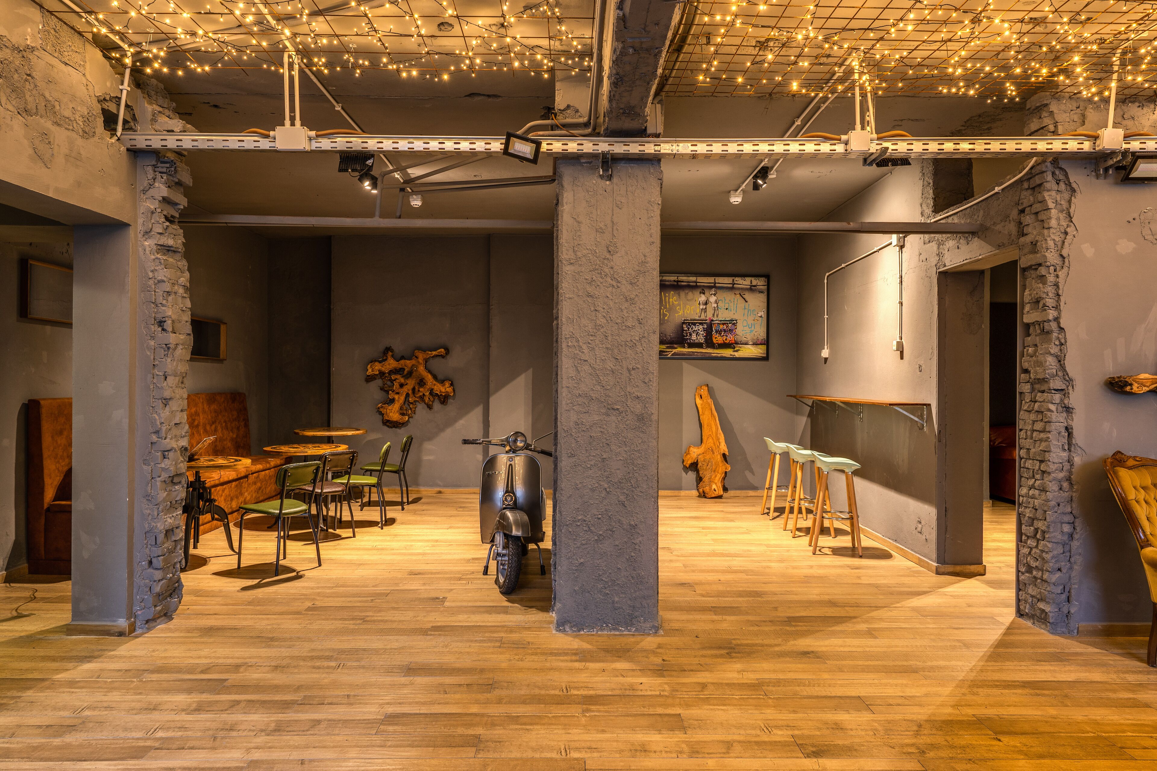 Interior com paredes de tijolo aparente, piso de madeira e uma scooter. Há mesas e cadeiras, e luzes de Natal no teto.