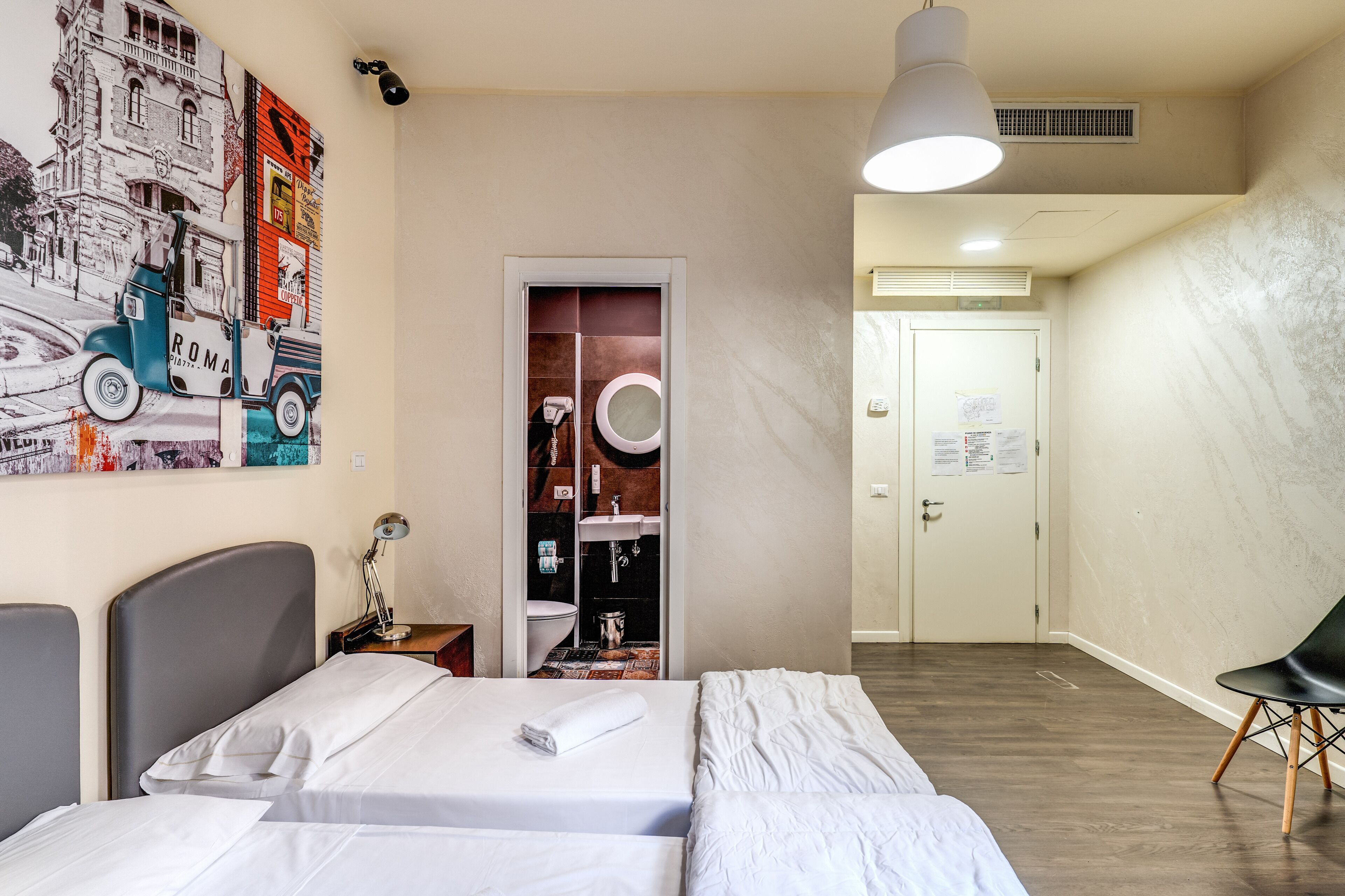 Habitación de hotel con dos camas, baño con inodoro y lavamanos, un cuadro en la pared y una puerta que da a otra habitación.