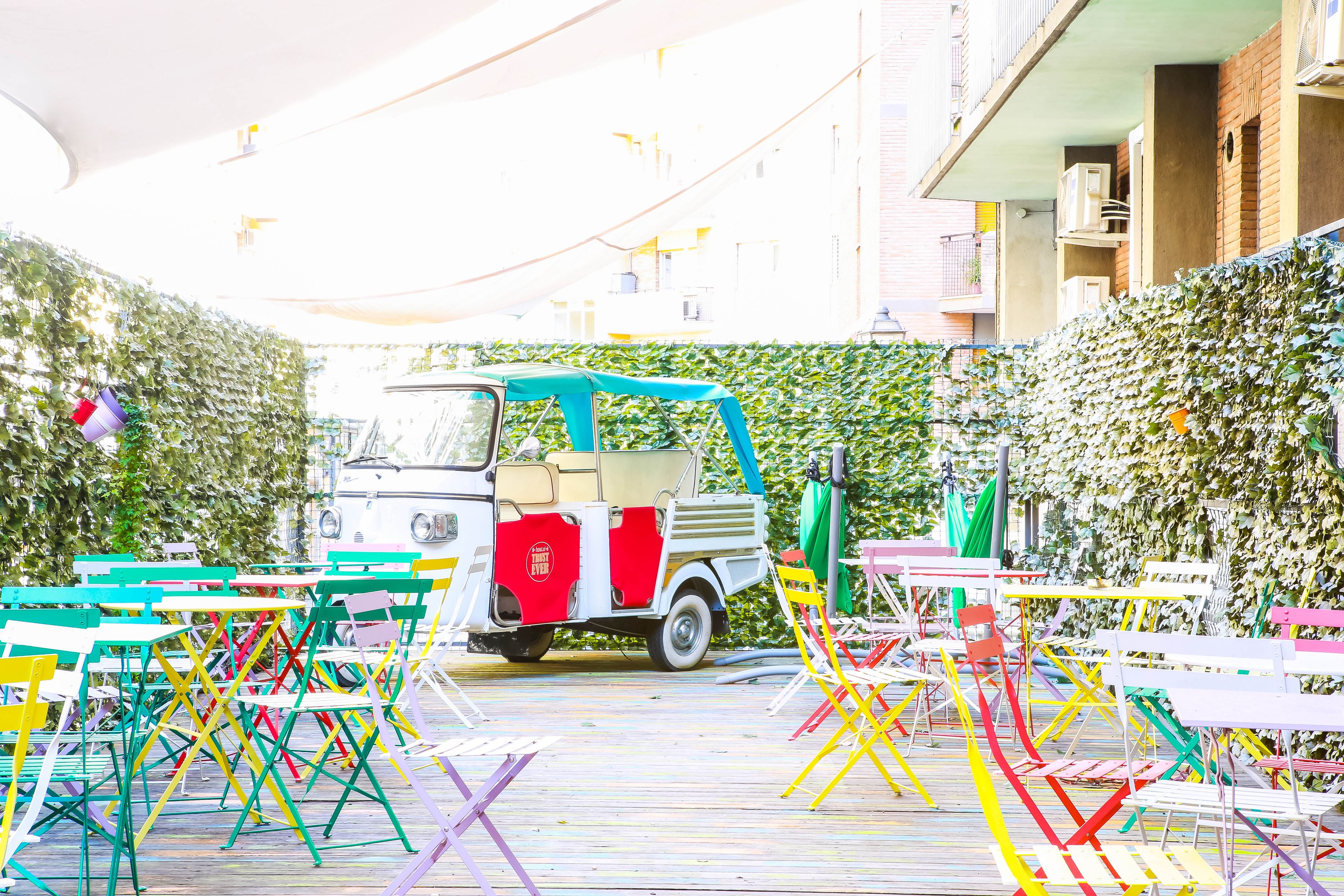 Un patio con sillas plegables y mesas de colores, un vehículo pequeño y una pared de seto.