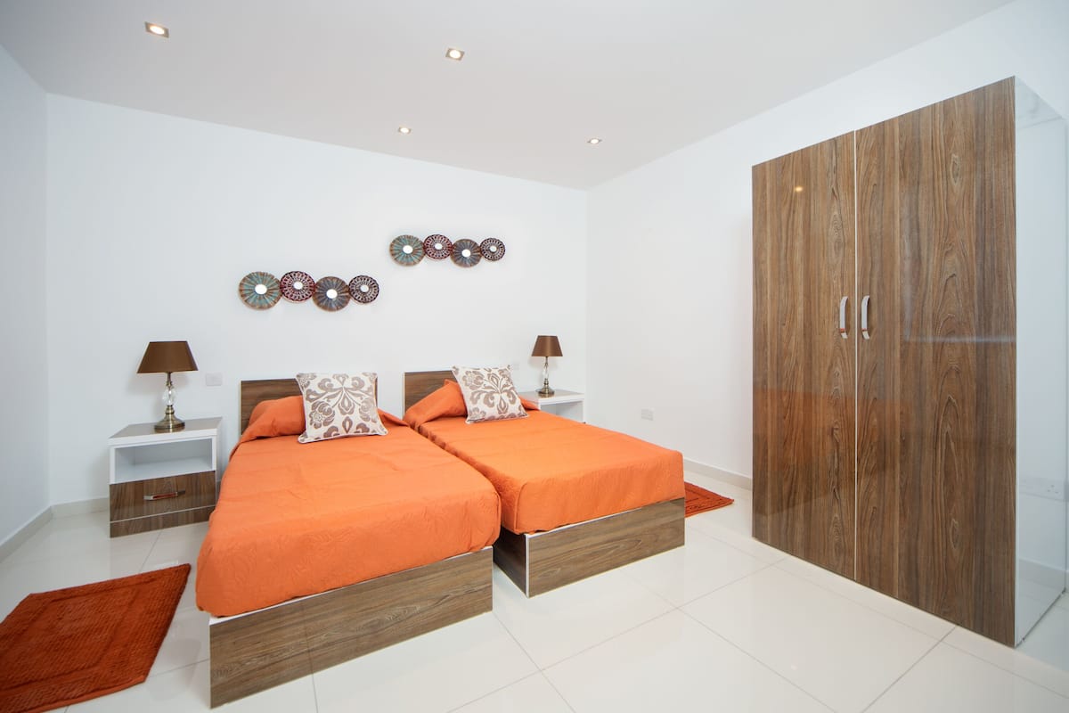 A modern bedroom with two beds, a wardrobe, decorative wall art, and a nightstand with a lamp.