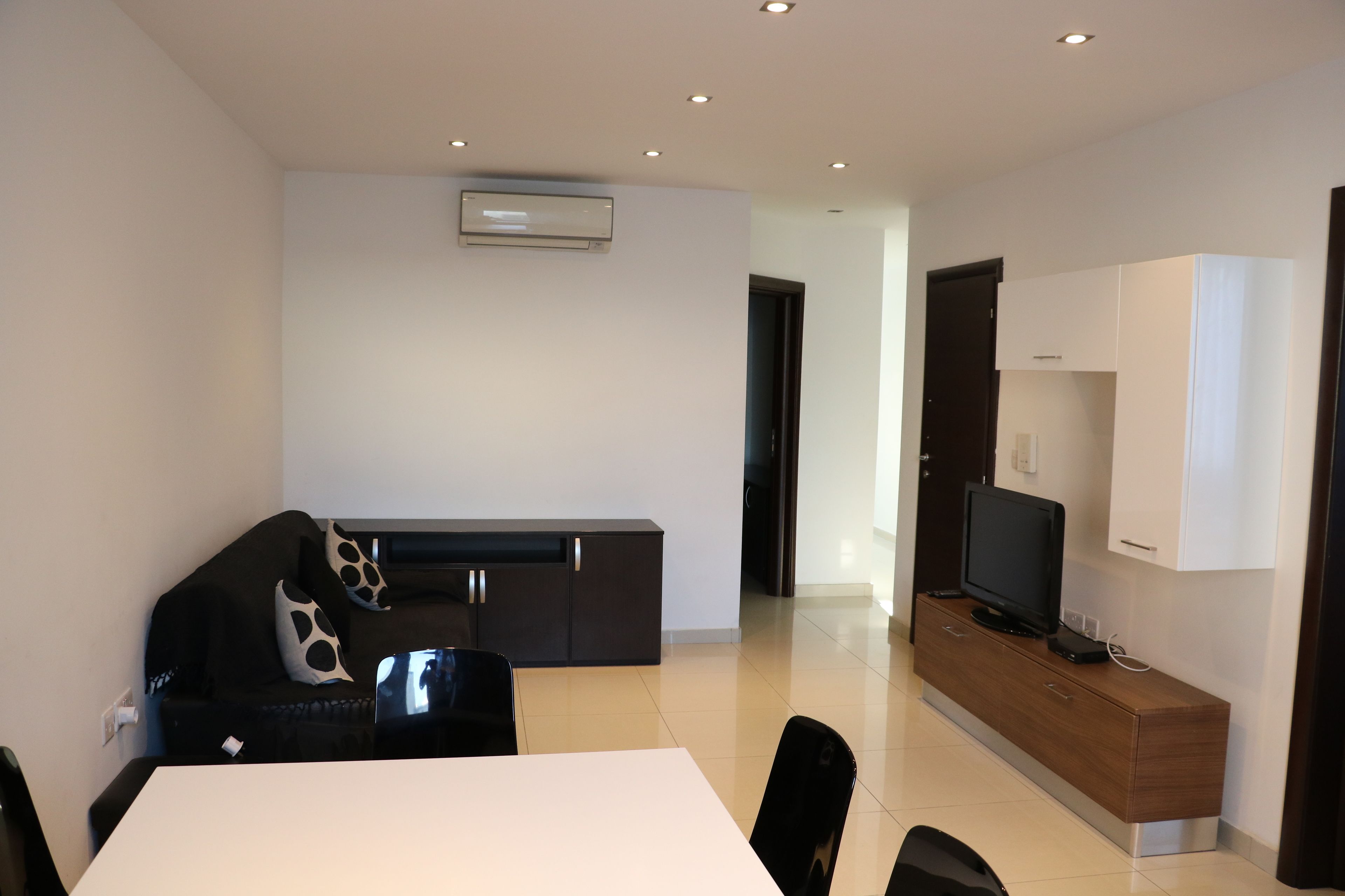 A modern living room with a dining table, black chairs, a sofa with patterned cushions, a TV on a wooden cabinet, and an air conditioning unit on the wall.