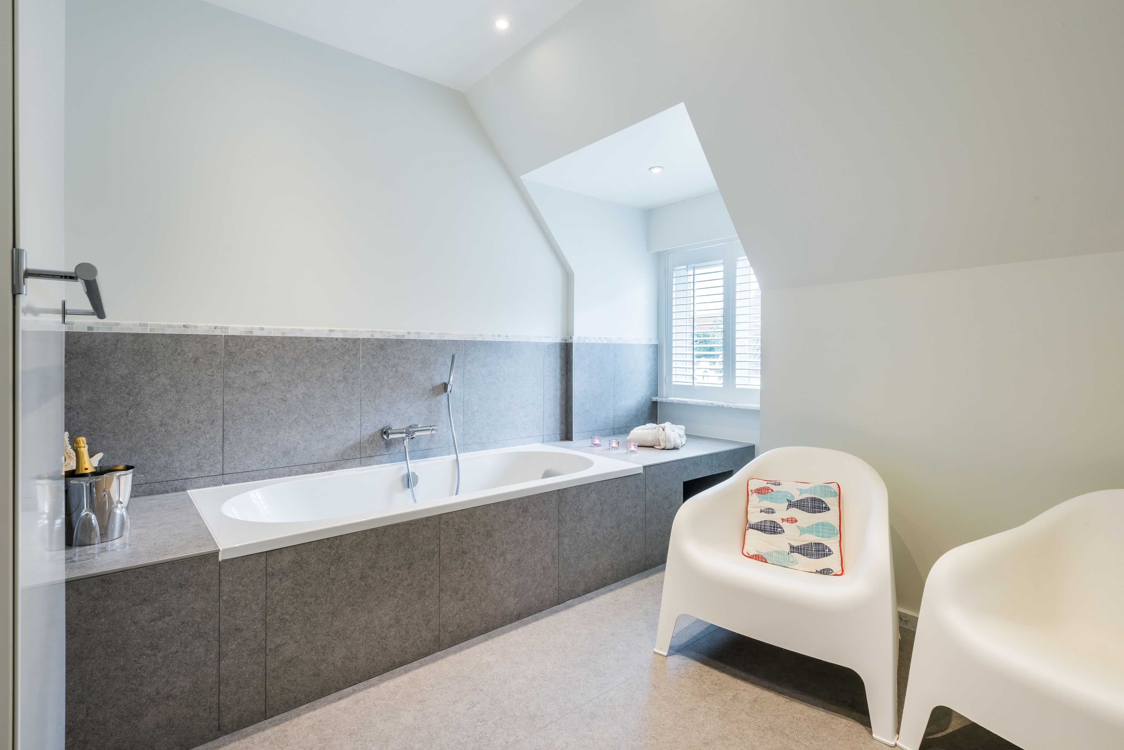 A modern bathroom with a large bathtub, a sink, and a window. There are two white chairs and a towel rack.