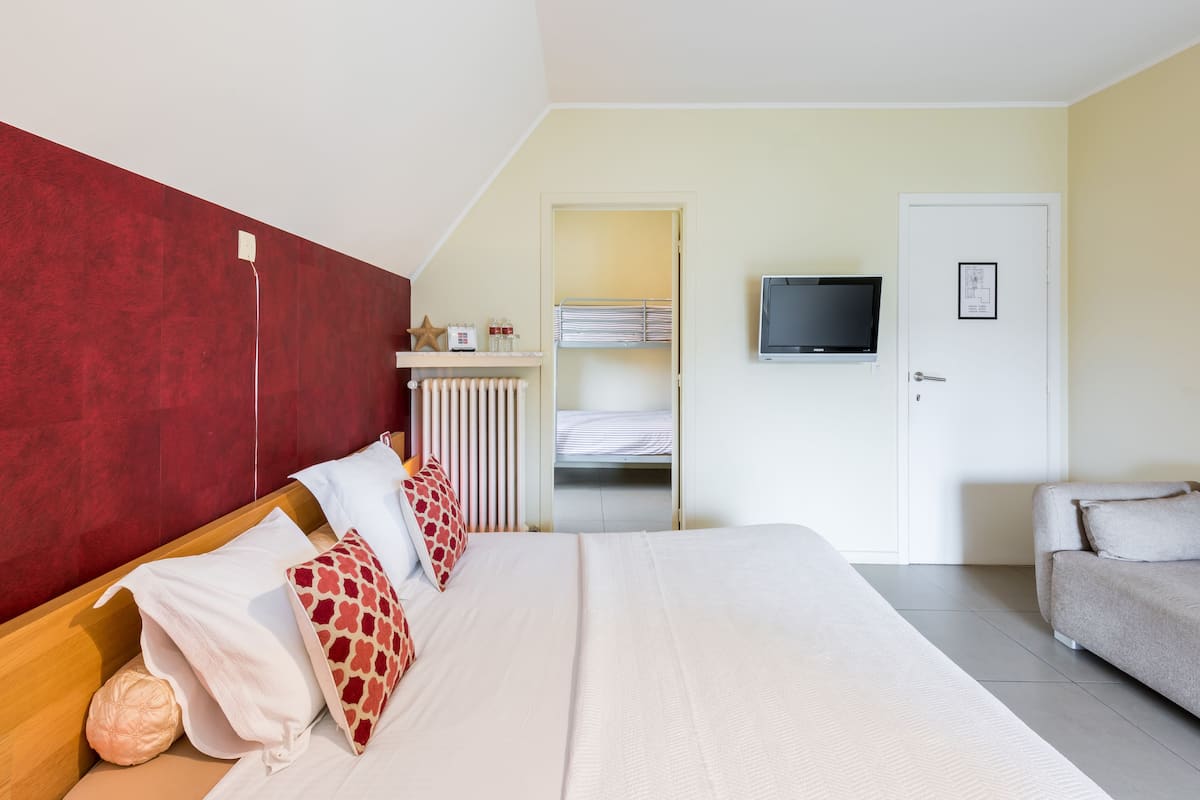 A hotel room with a large bed, a TV mounted on the wall, a red accent wall, a white door, and a grey sofa.