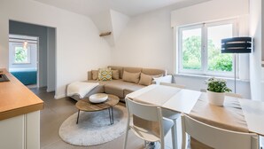 A modern, well-lit living room with a sofa, coffee table, and dining area. A large window provides natural light and a view of greenery outside. - B&B Vis-à-Vis (De Haan)