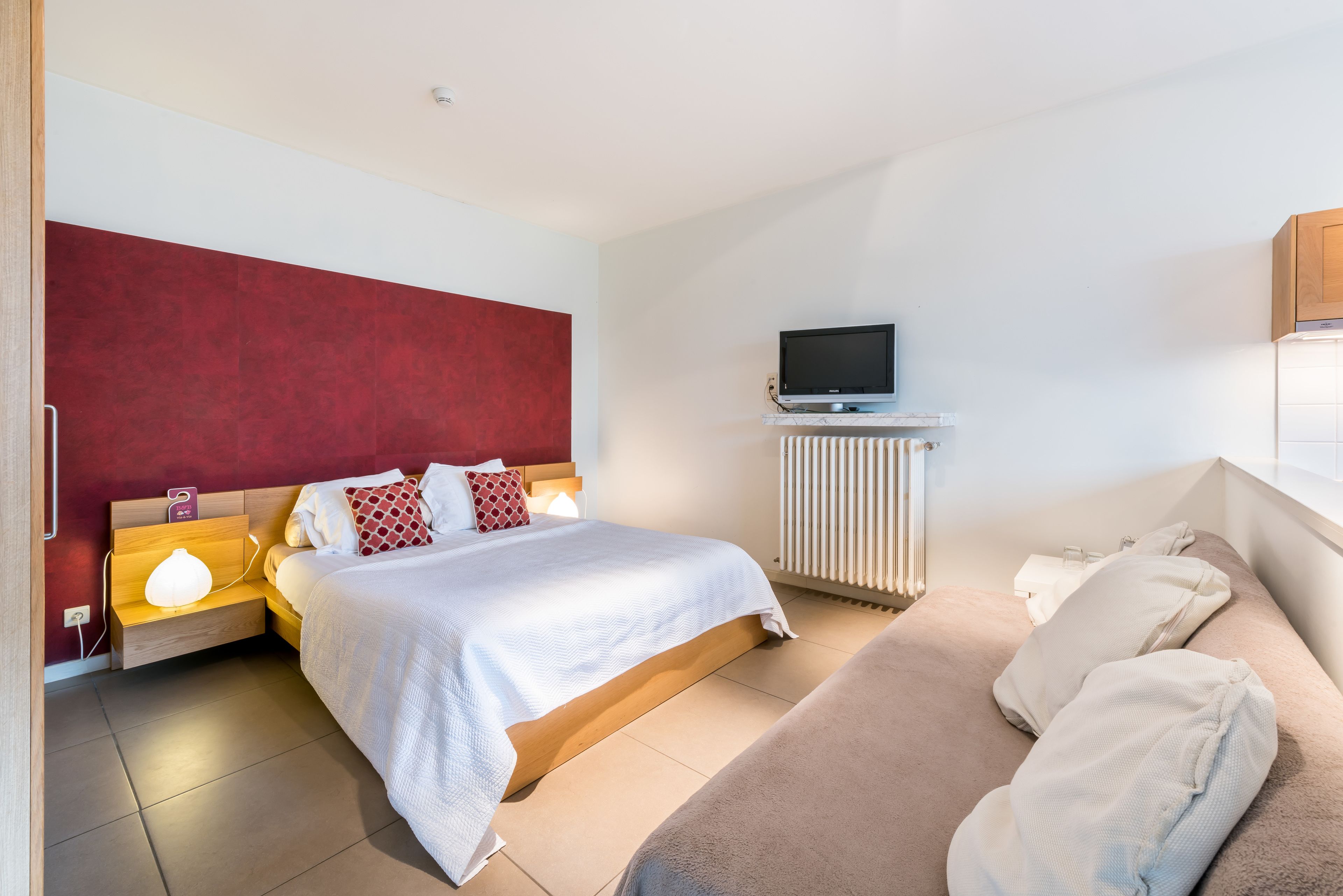 A modern hotel room with a large bed, a sofa, a TV on a stand, and a bedside table with a lamp.