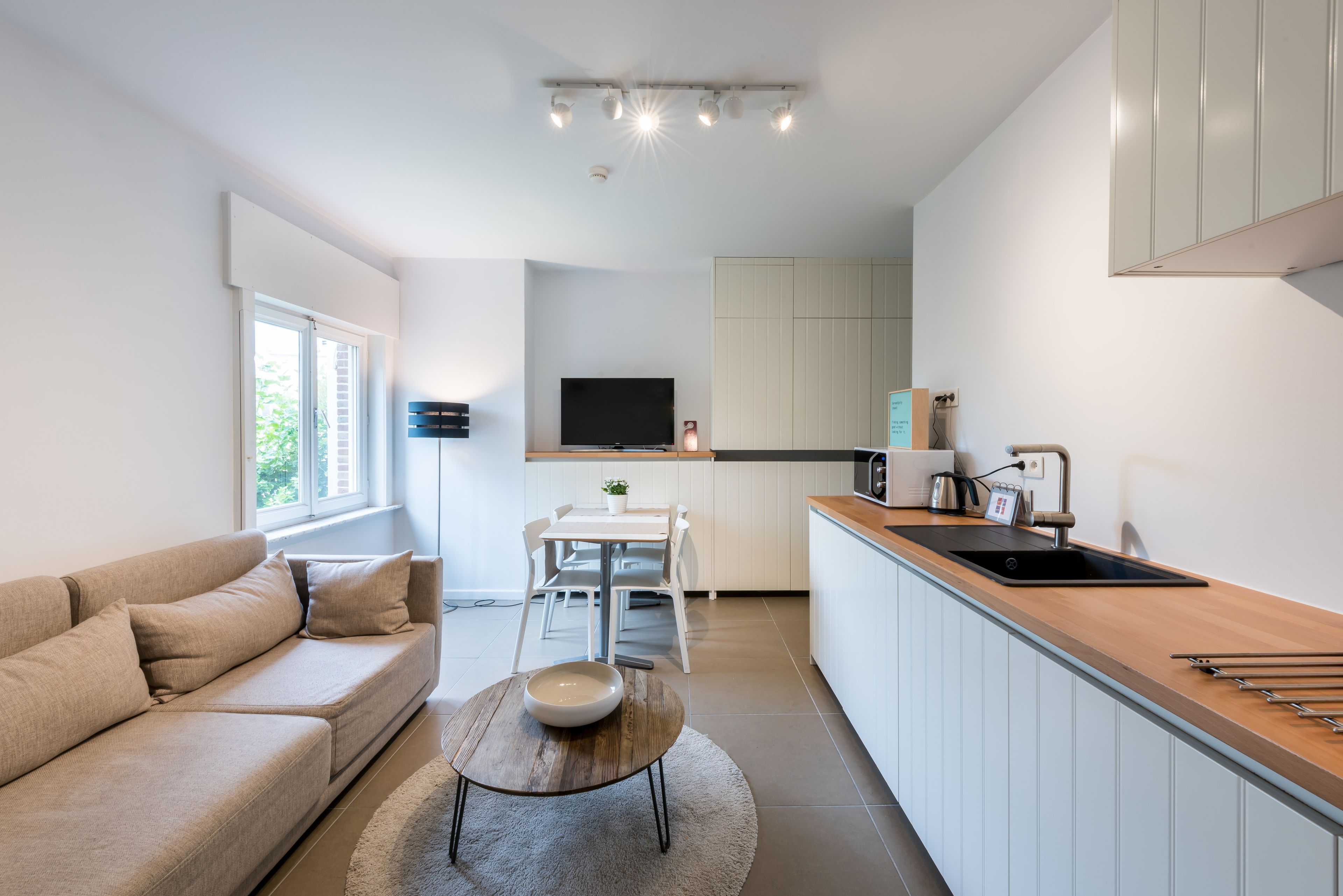 Een moderne keuken met witte kastjes, een houten aanrechtblad en een inbouwoven. Er is een eethoek met een witte tafel en stoelen, en een bank in de woonkamer.
