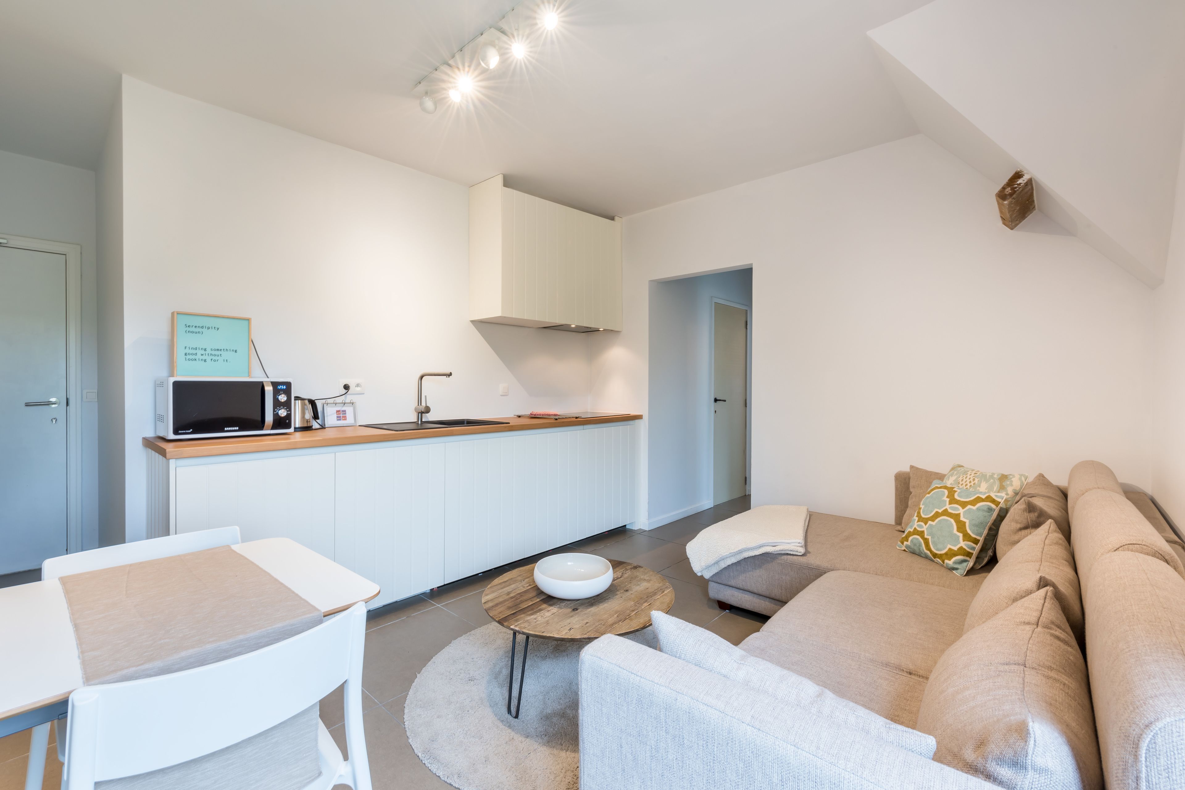 A modern living room with a sofa, coffee table, and a small kitchen area with a microwave and a sink.