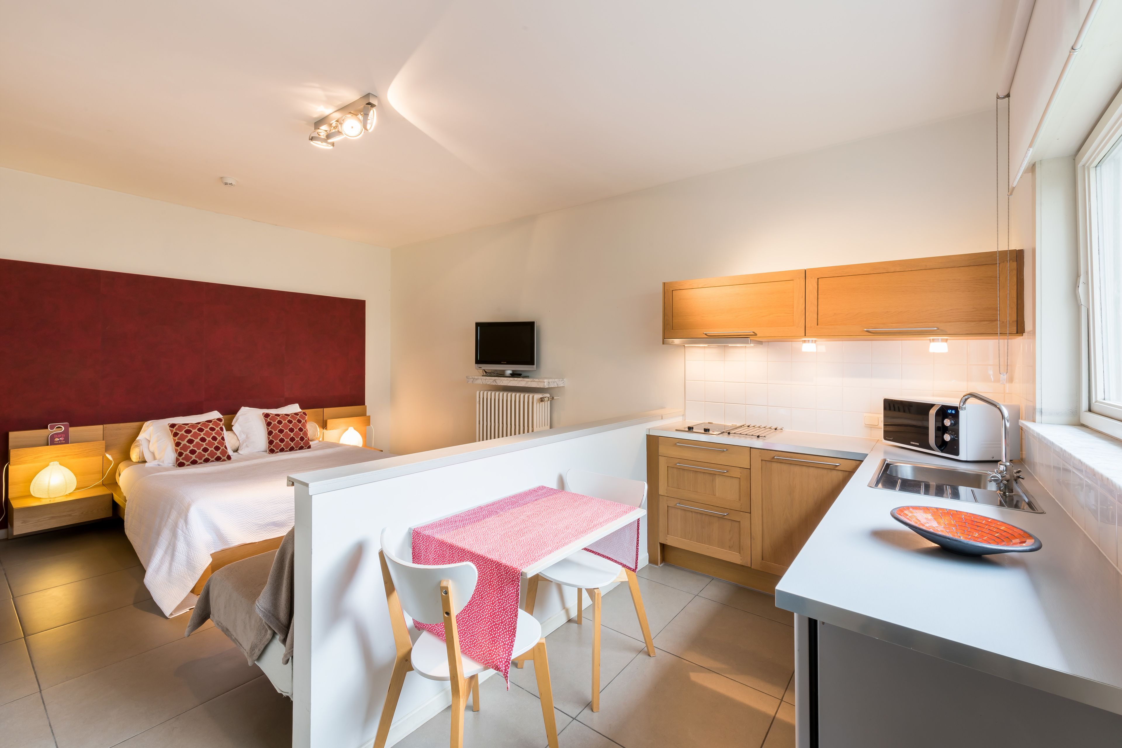 Habitación de hotel compacta con kitchenette, cama con pared de acento roja, televisor y zona de comedor con mesa y sillas.