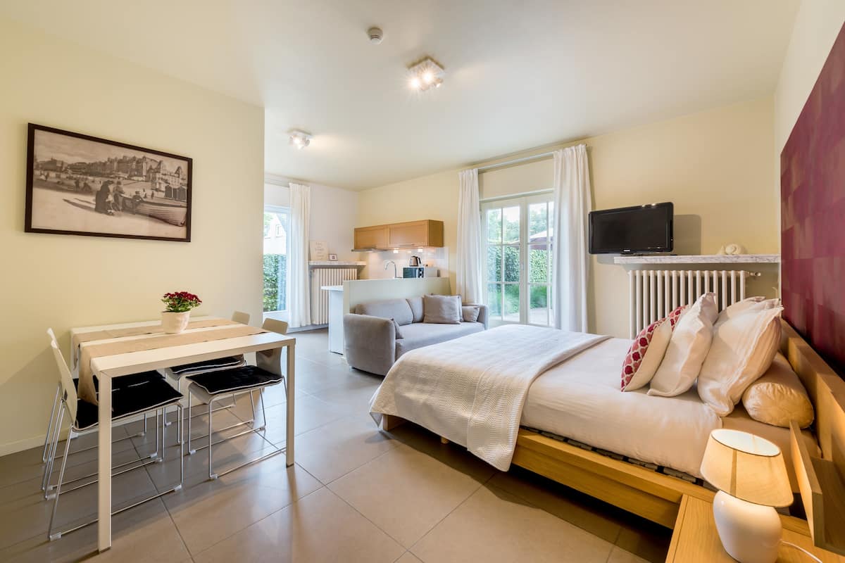 A modern hotel room with a bed, bedside table, lamp, and a dining area with chairs and a table.