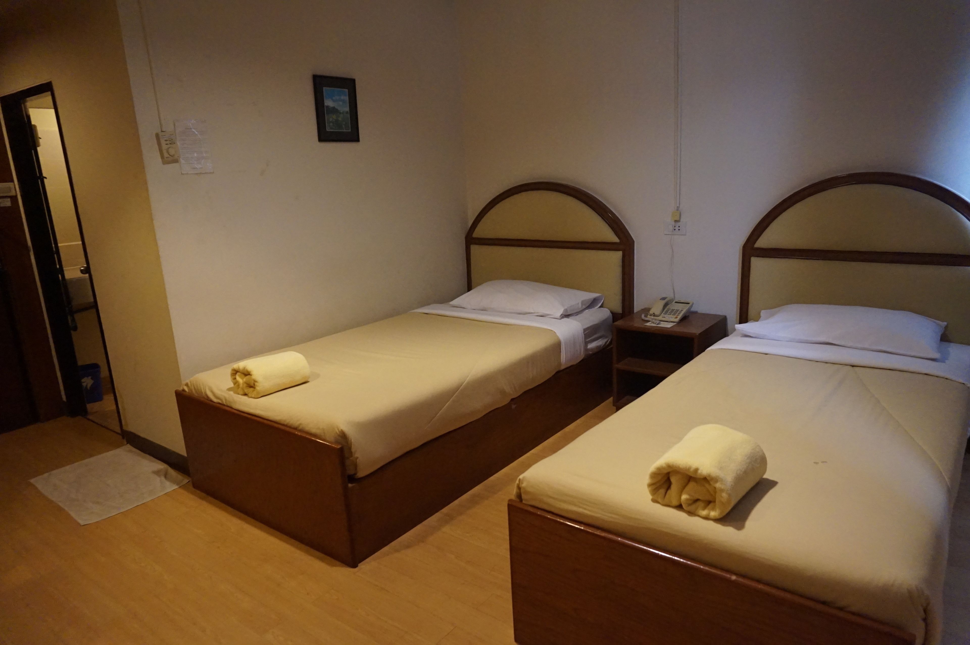 Dos camas individuales con cabeceros de madera y ropa de cama blanca, una mesita de noche con un teléfono, y un pequeño cuadro colgado en la pared.
