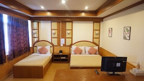 Habitación de hotel con dos camas individuales, un escritorio de madera, un televisor y una pequeña estantería con un teléfono y papeles.