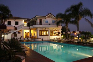 Un edificio de dos pisos con piscina, sillas de descanso y sombrillas.