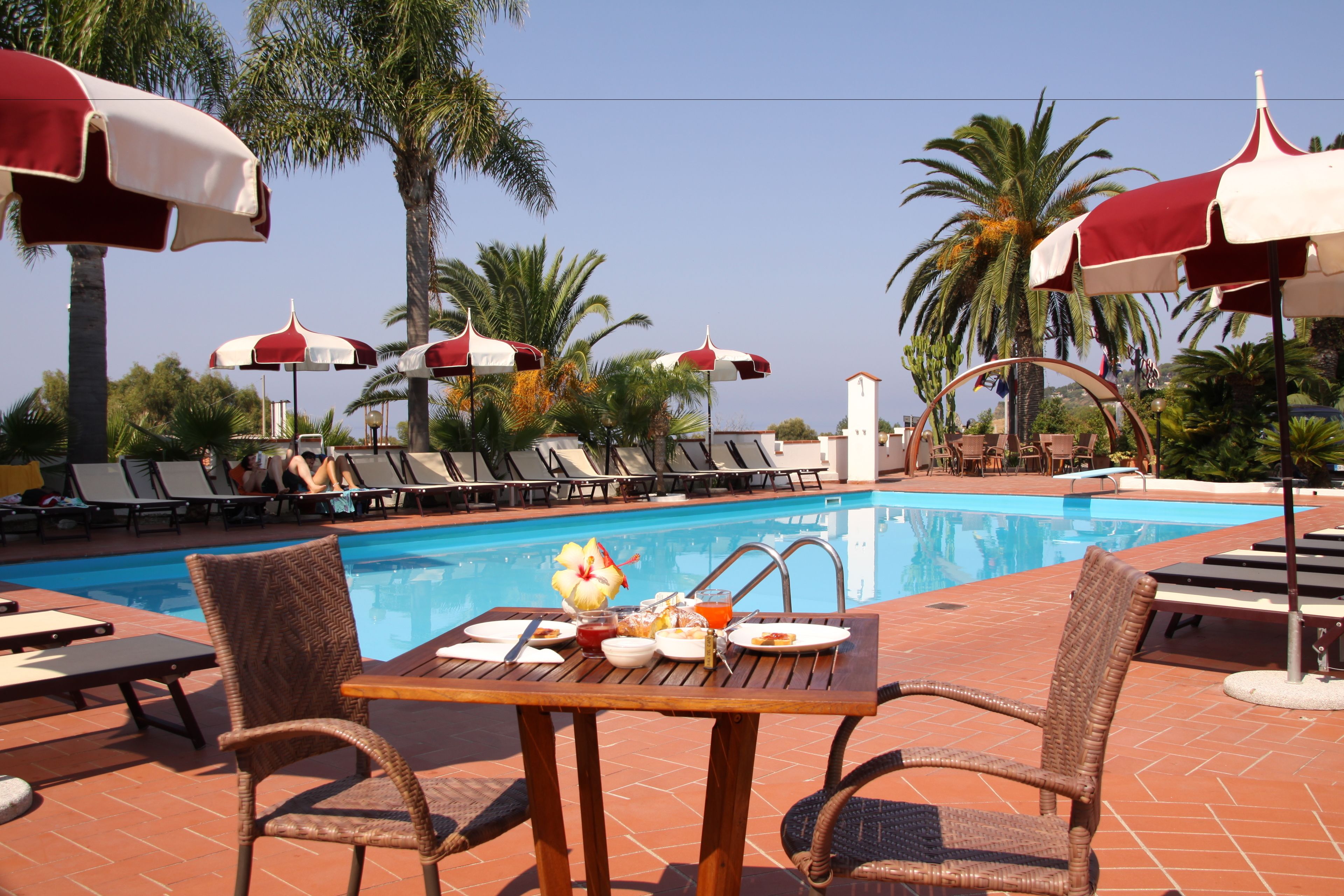 A poolside area with lounge chairs and umbrellas, a dining table with breakfast items, and a clear blue pool.