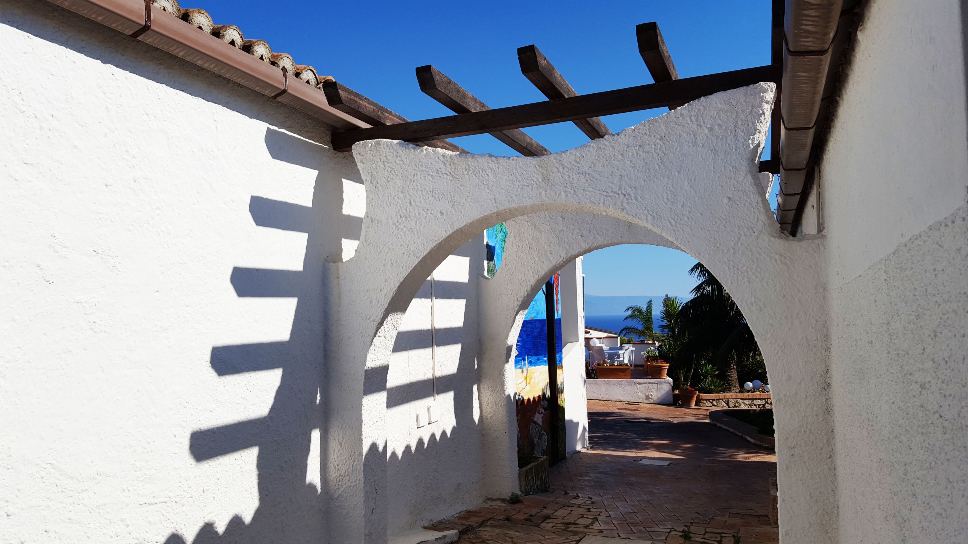 Ein rundbogiger Durchgang führt zu einem Innenhof mit Fliesenboden, einer Pergola und Blick aufs Meer.