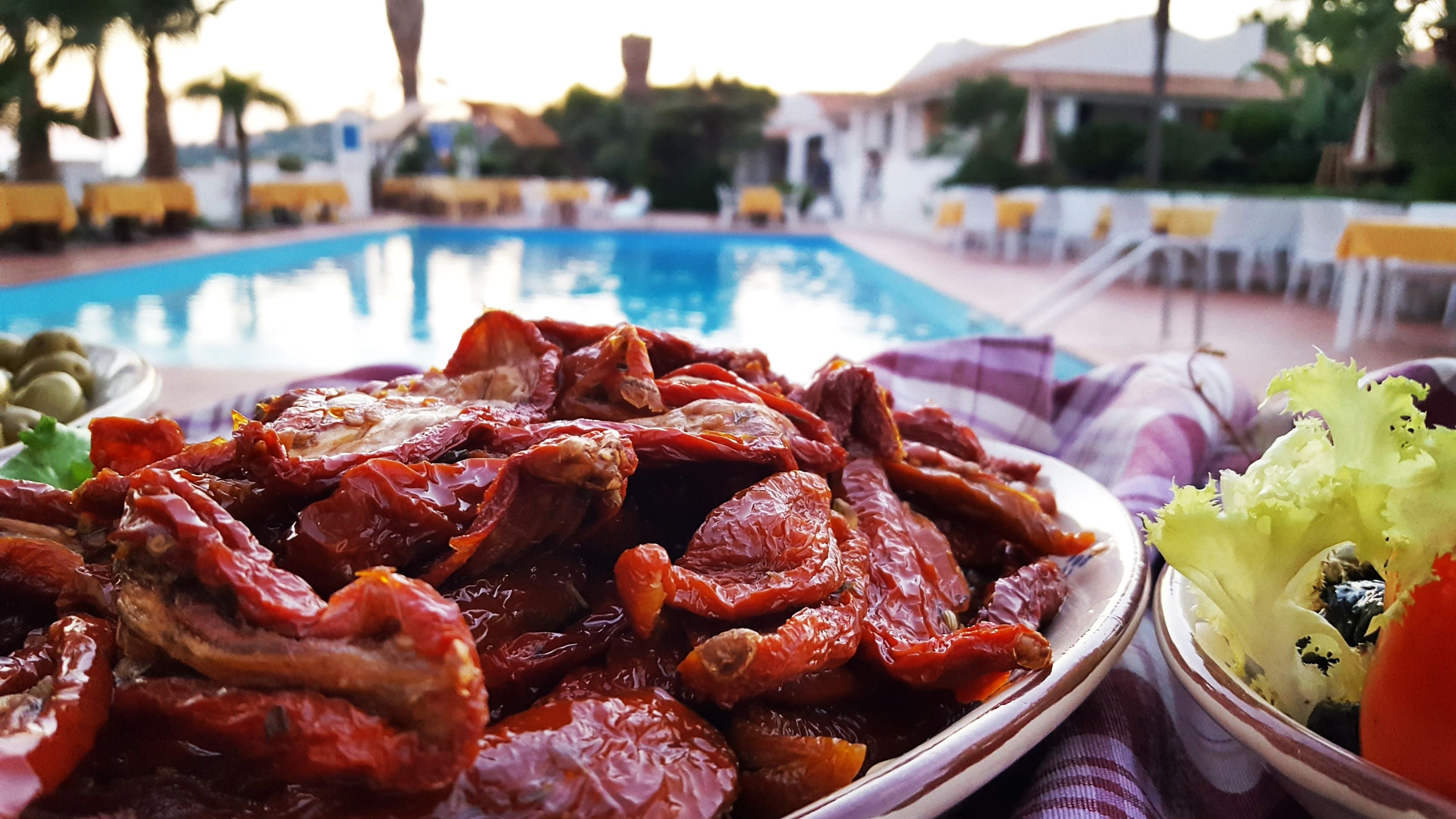 A dining table with a plate of roasted meat and vegetables, a bowl of salad, and a pool in the background.