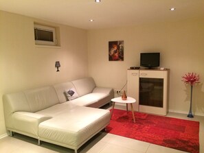 A modern living room with a white sofa, a small white table, a red rug, a television on a stand, and a vase with a red flower. - Ferienhaus Piešťany (Banka)