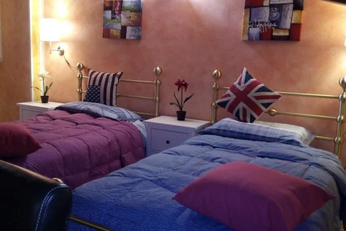 A hotel room with two beds, each with a different color bedspread, a bedside table with a lamp, and a wall with a painting.