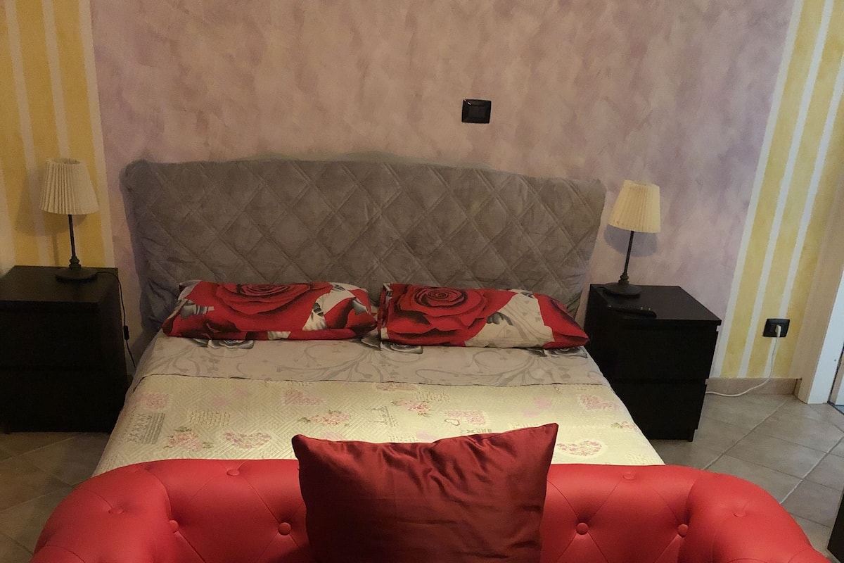 A bedroom with a bed, a red upholstered sofa, two nightstands, and a wall-mounted light switch.