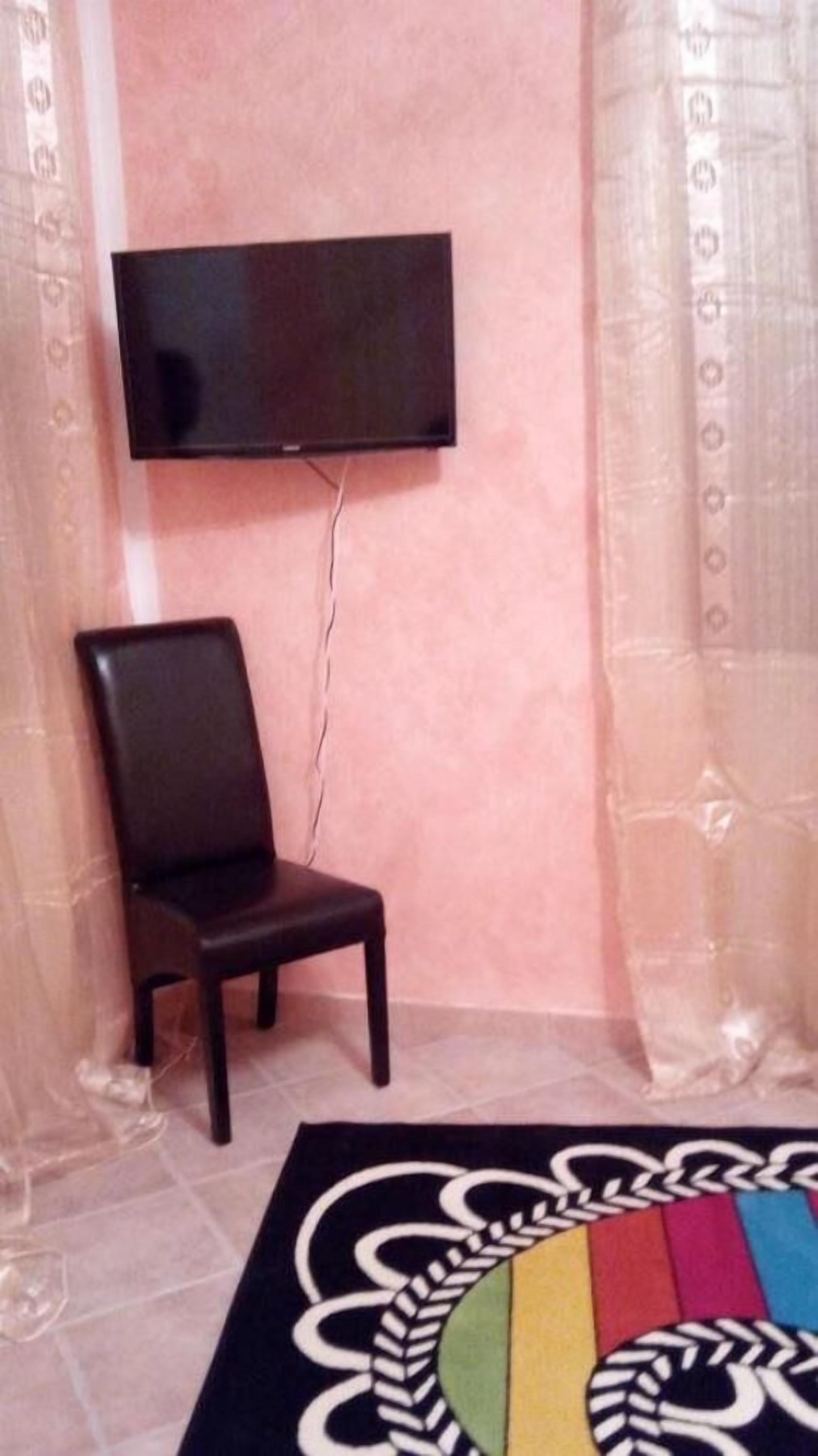 Ein Zimmer mit einem an der Wand befestigten Flachbildfernseher, einem dunklen Holzstuhl und einem farbenfrohen Teppich auf dem Boden.