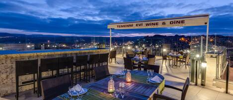 An outdoor dining area with tables set for two, a view of the cityscape, and a sign that reads "TEMENNİ EVI WINE & DINE".