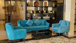 A lobby with a blue sofa set, a coffee table, and a decorative glass display case with various items. - TTC Hotel – Hoi An (Da Nang)
