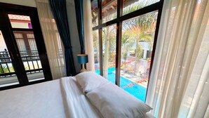 A hotel room with a bed, a window with curtains, and a view of a swimming pool and outdoor area. - TTC Hotel – Hoi An (Da Nang)